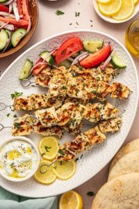 Overhead shot of a serving platter with grilled chicken souvlaki skewers, tzatziki sauce, lemon slices, pita bread, and a fresh tomato-cucumber salad.