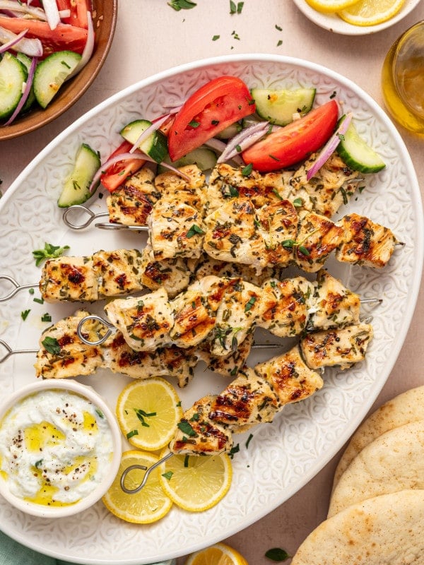 Overhead shot of a serving platter with grilled chicken souvlaki skewers, tzatziki sauce, lemon slices, pita bread, and a fresh tomato-cucumber salad.