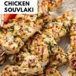 Side view of juicy chicken souvlaki skewers on a white patterned platter with cucumber tomato salad and a small bowl of tzatziki with text overlay that reads " Greek Chicken Souvlaki"