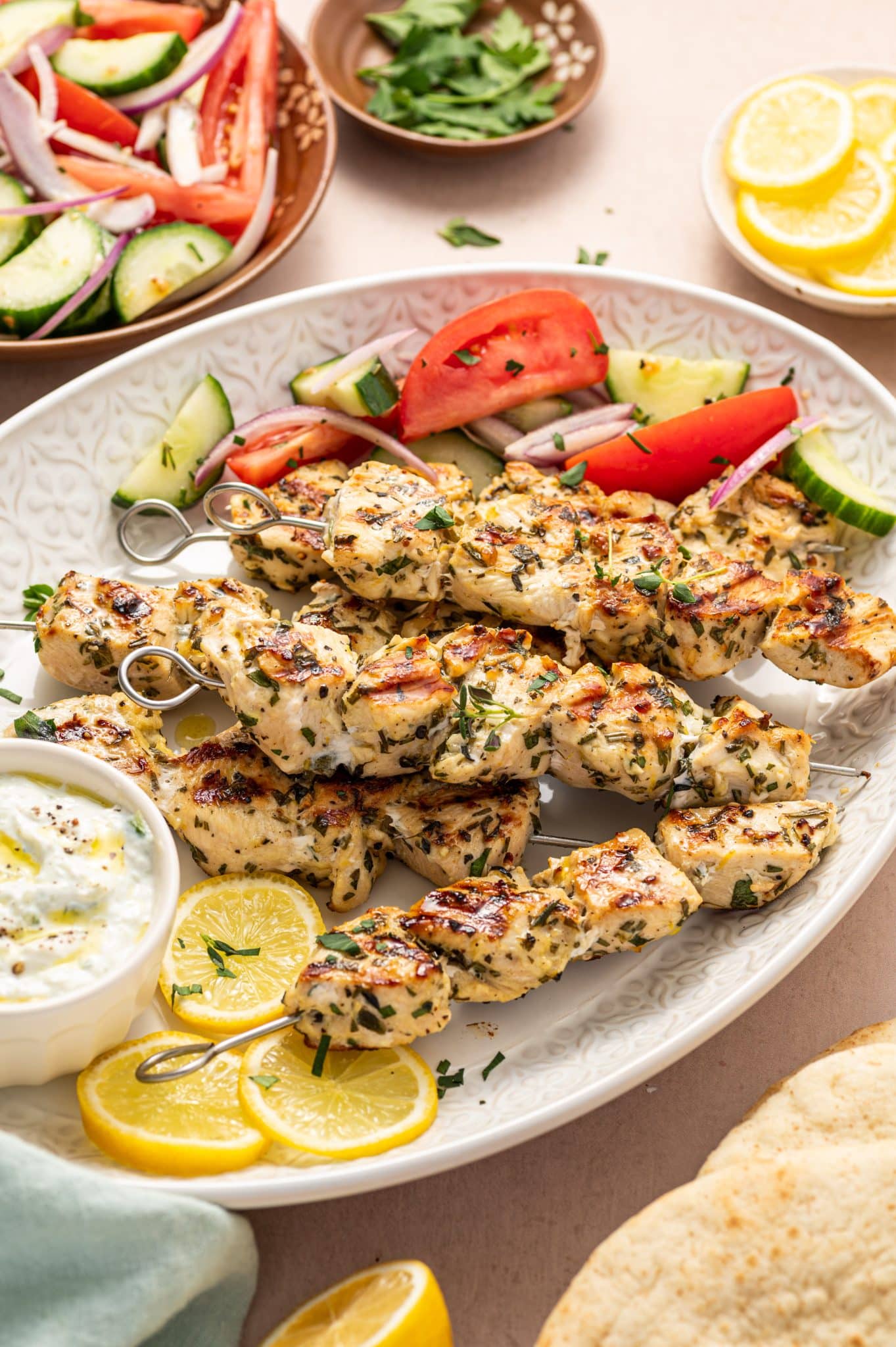 Angled view of a platter loaded with grilled chicken souvlaki skewers, lemon slices, tzatziki, and Greek salad with tomatoes, cucumbers, and red onions.