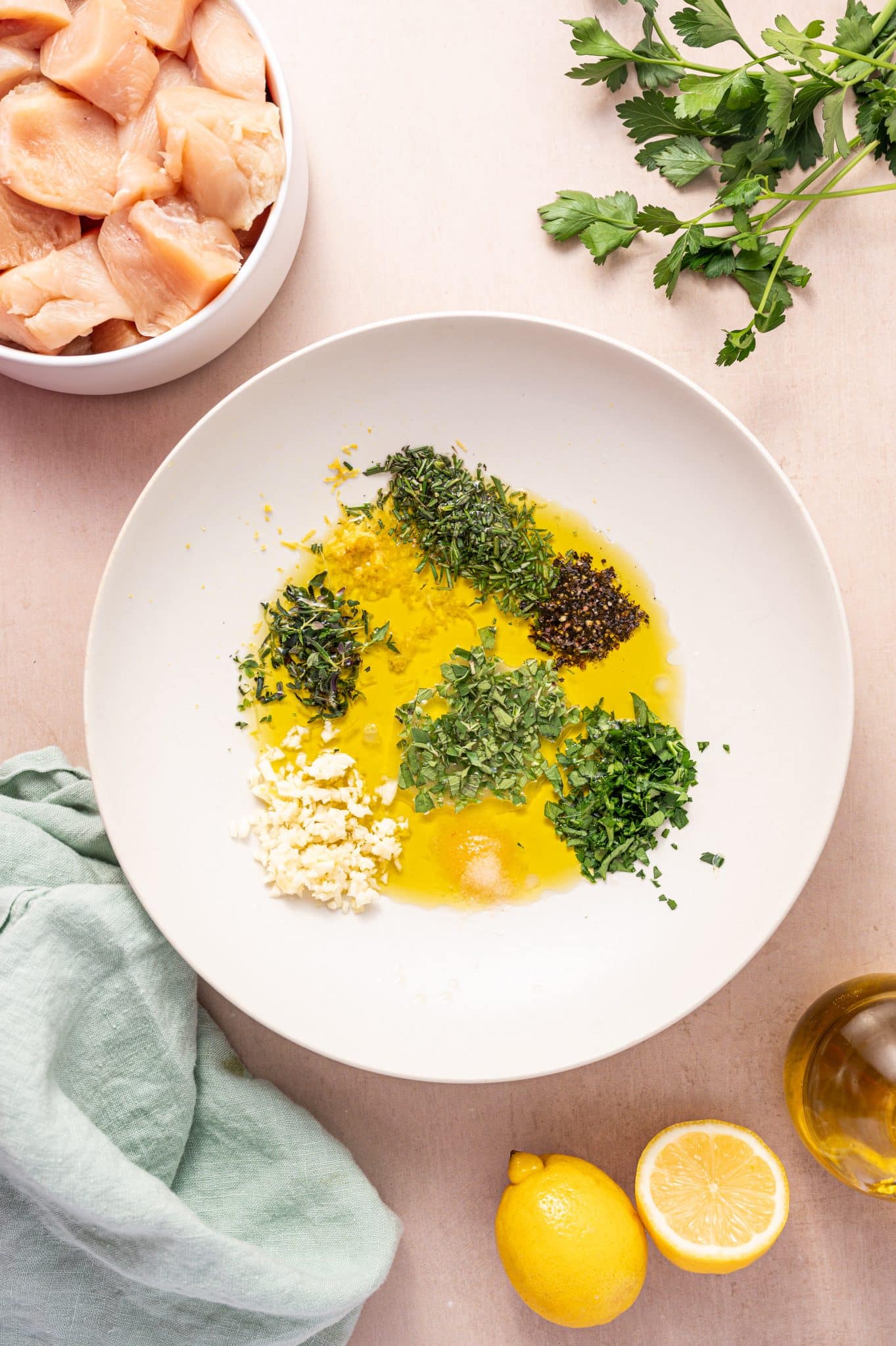 Flat lay of a white bowl filled with olive oil, minced garlic, fresh chopped herbs, lemon zest, and black pepper ready to be mixed into a Greek chicken souvlaki marinade. Surrounding ingredients include raw chicken cubes, lemons, fresh parsley, and a bottle of olive oil.