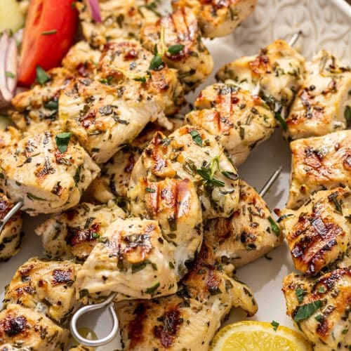 Side view of juicy chicken souvlaki skewers on a white patterned platter with cucumber tomato salad and a small bowl of tzatziki.