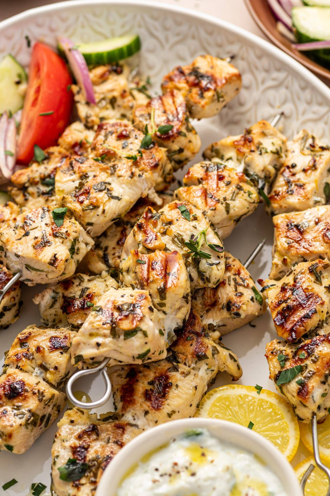 Side view of juicy chicken souvlaki skewers on a white patterned platter with cucumber tomato salad and a small bowl of tzatziki.