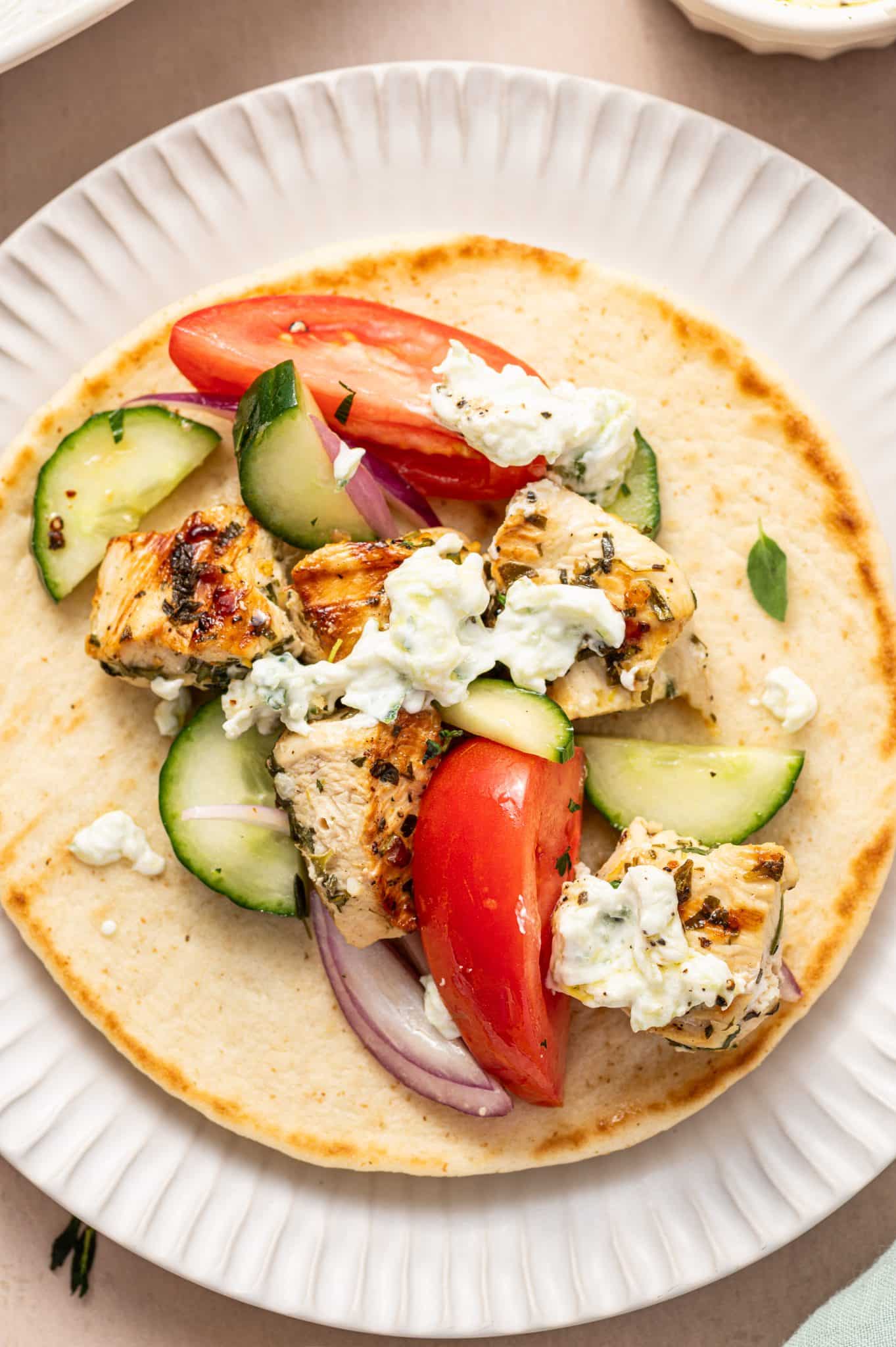 Overhead view of an open pita topped with grilled chicken souvlaki, fresh cucumbers, tomatoes, red onions, and a drizzle of tzatziki sauce.
