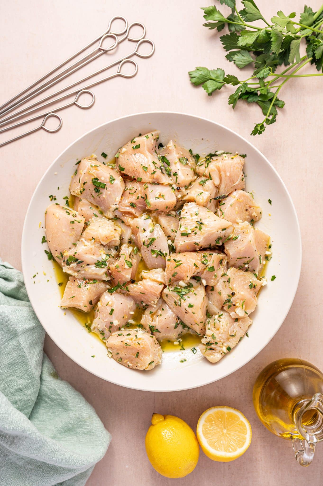 Chunks of raw chicken marinating in a lemon herb and olive oil mixture in a white bowl, surrounded by skewers, fresh parsley, and whole lemons—ready to be made into Greek souvlaki.