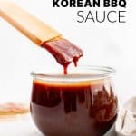 A vertical image of thick Korean BBQ sauce dripping off a wooden brush into a small glass jar, styled on a neutral background with bold text overlay reading “Korean BBQ Sauce.”