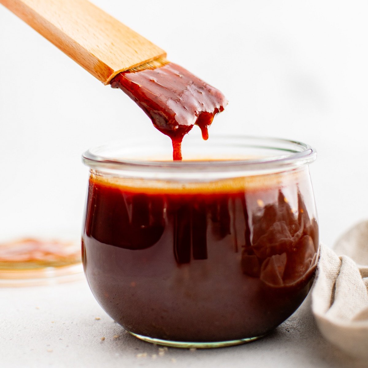A wooden basting brush drips thick Korean BBQ sauce back into a glass jar. The sauce is deep red and glossy, showing its ideal sticky consistency.