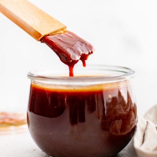 A wooden basting brush drips thick Korean BBQ sauce back into a glass jar. The sauce is deep red and glossy, showing its ideal sticky consistency.