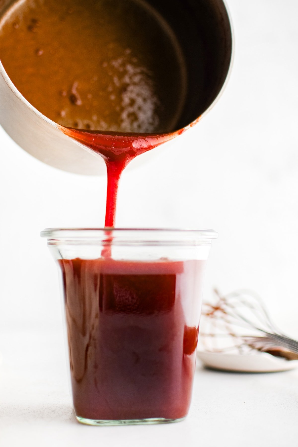A close-up of deep red mumbo sauce being poured from a saucepan into a clear glass storage jar.
