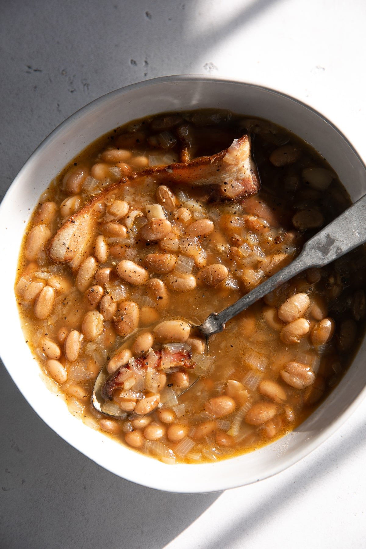 Hearty pinto beans simmered with bacon and onions, served in a white bowl with a rich, smoky broth and a spoon for serving.