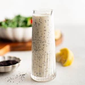 A minimalist shot of a tall glass filled with creamy poppyseed dressing, with salad greens and lemon in the soft-focus background.