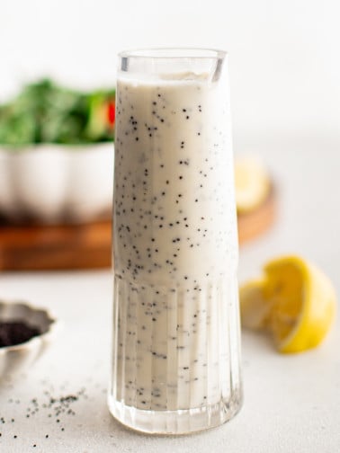 A minimalist shot of a tall glass filled with creamy poppyseed dressing, with salad greens and lemon in the soft-focus background.