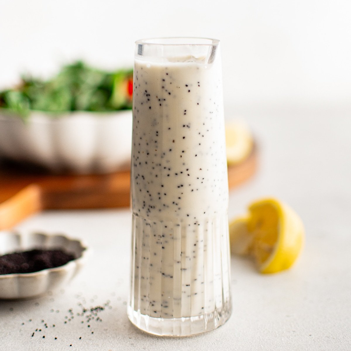 A minimalist shot of a tall glass filled with creamy poppyseed dressing, with salad greens and lemon in the soft-focus background.