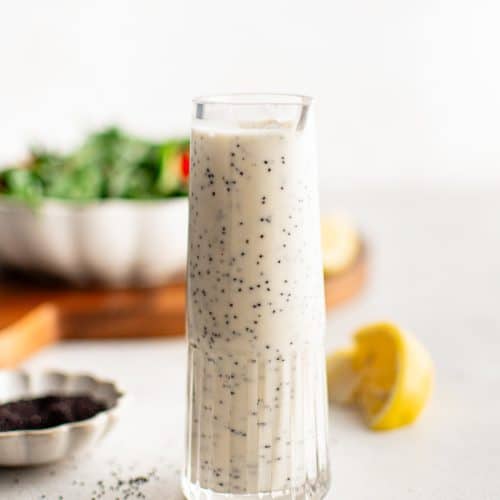 A minimalist shot of a tall glass filled with creamy poppyseed dressing, with salad greens and lemon in the soft-focus background.