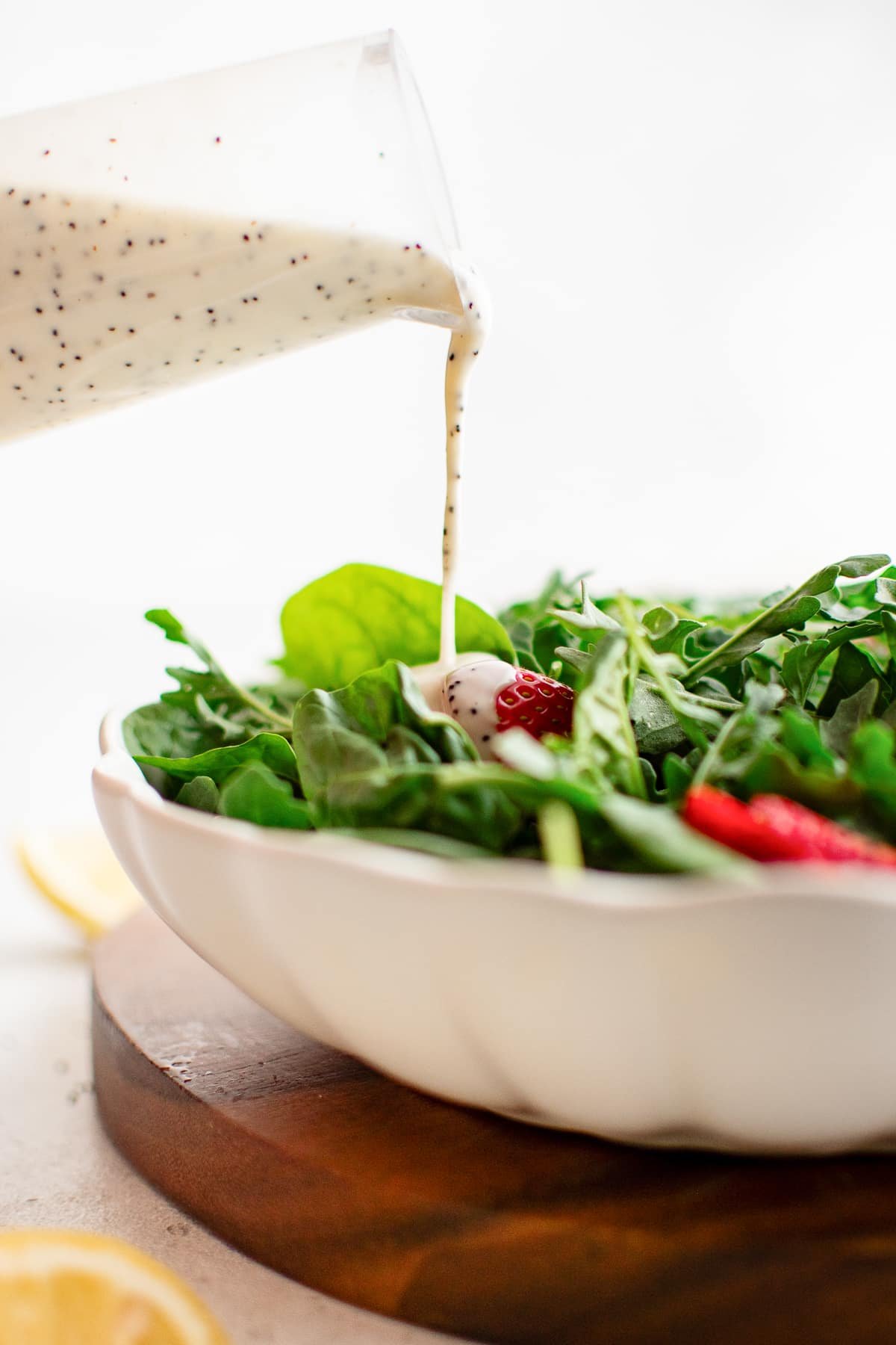 A glass container pours creamy poppyseed dressing over a fresh spinach salad with sliced strawberries.