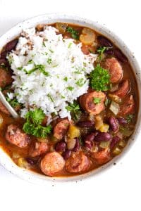 A hearty bowl of red beans stewed with smoked sausage, bell peppers, and onions, served alongside a generous scoop of fluffy white rice and garnished with parsley.