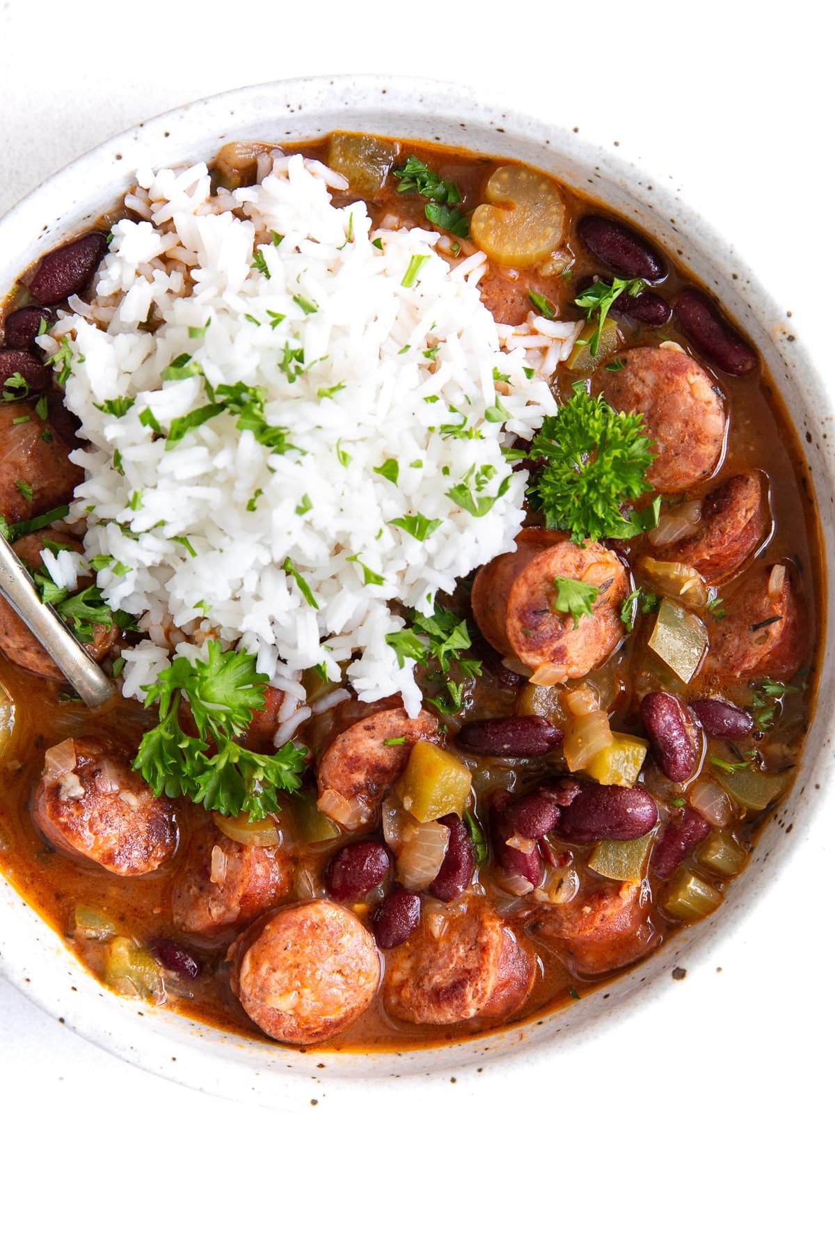 A hearty bowl of red beans stewed with smoked sausage, bell peppers, and onions, served alongside a generous scoop of fluffy white rice and garnished with parsley.