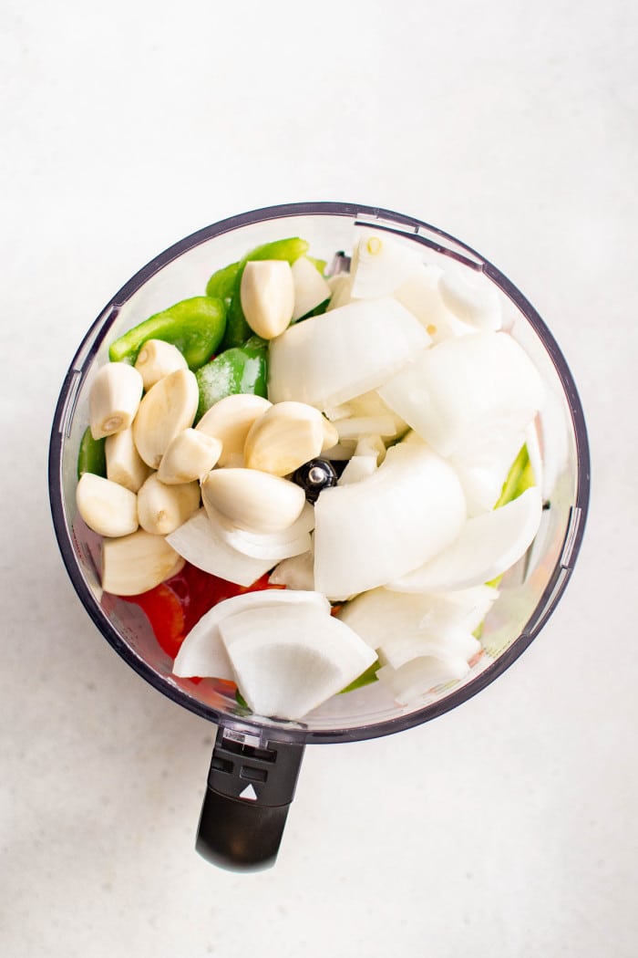 A top-down view of chopped garlic, white onion, sweet peppers, and green bell peppers in a food processor, ready to be blended into sofrito.