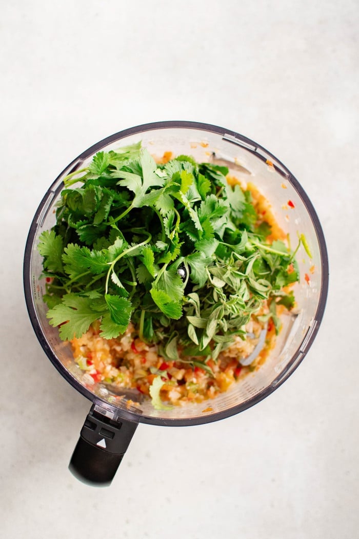 Fresh cilantro and oregano added to partially blended sofrito in a food processor