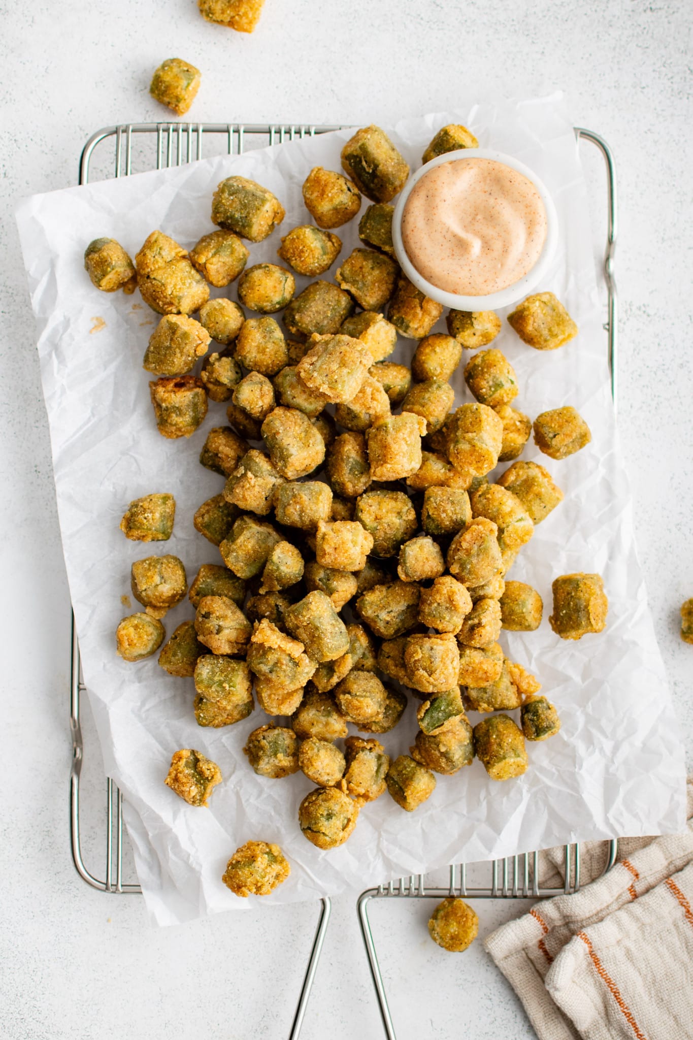 A batch of golden brown fried okra laid out on parchment-lined wire rack with a side of remoulade-style dipping sauce. The okra pieces are evenly coated in a cornmeal crust.