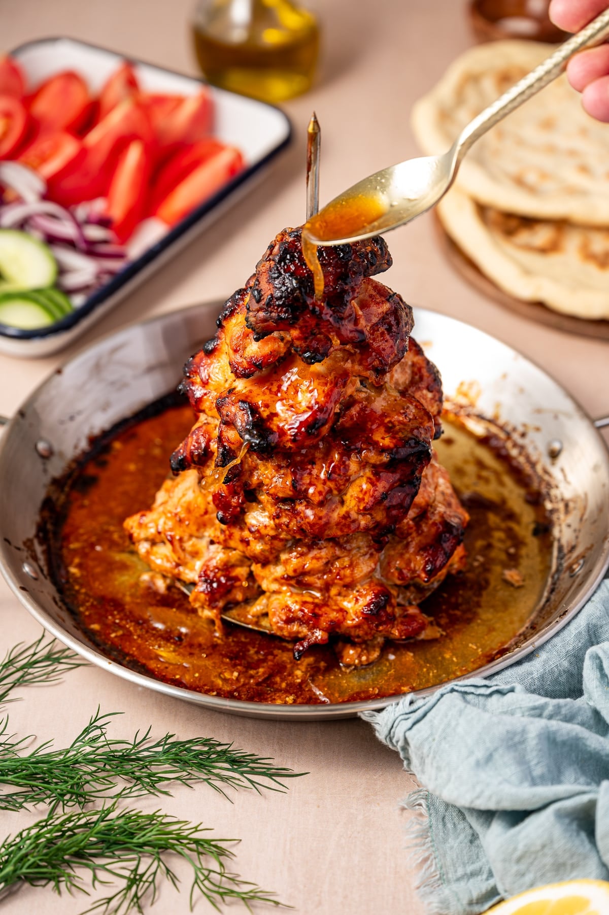 Close-up of golden roasted chicken gyro meat on a vertical spit being basted with pan juices from a silver spoon, with charred edges, a silver pan, sliced tomatoes, red onion, cucumber, warm pita bread, and olive oil in the background.