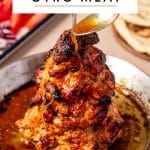 Pinterest pin image showing homemade chicken gyro meat stacked on a vertical spit in a silver pan with golden pan juices, dark charred edges, and a spoon drizzling juices over the top. A white text box at the top reads โDIY CHICKEN GYRO MEATโ in large black letters, and โTHEFORKEDSPOON.COMโ appears at the bottom. No garnish is on the meat, with fresh vegetables and pita bread blurred in the background.