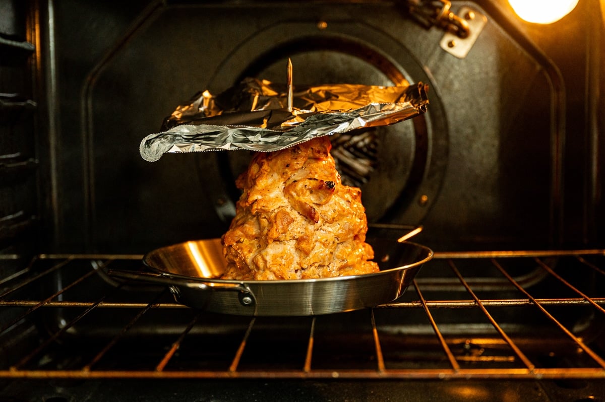 Chicken gyro meat roasting upright on a vertical spit inside an oven, with the stacked marinated chicken sitting in a silver pan and covered loosely at the top with crinkled aluminum foil.