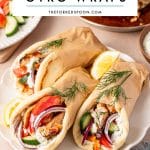 Pinterest pin image showing three chicken gyro wraps on a white scalloped plate, filled with sliced chicken gyro meat, white tzatziki sauce, red tomato slices, cucumber pieces, thin red onion slices, and fresh dill. The pitas are wrapped in brown parchment paper and served with lemon wedges, with a silver pan of chicken gyro meat, a small bowl of tzatziki, sliced vegetables, and a light blue napkin in the background. A white text box at the top reads โDIY CHICKEN GYRO WRAPSโ and โTHEFORKEDSPOON.COMโ in black text.