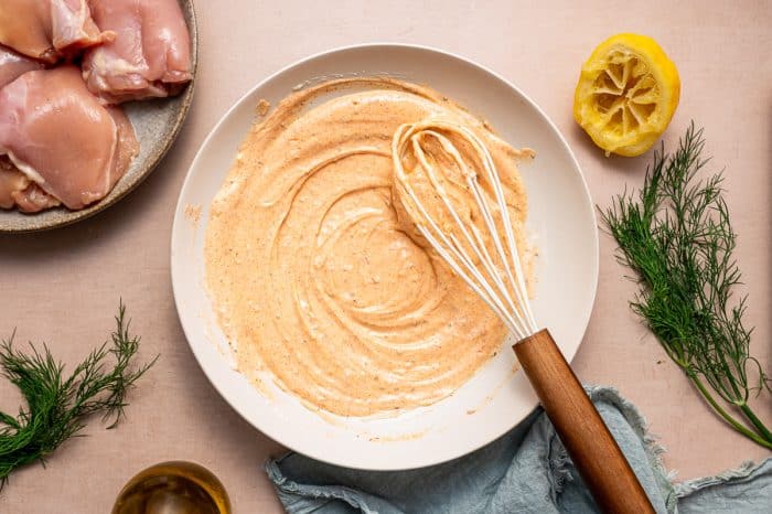 Overhead view of a white shallow bowl filled with creamy orange yogurt marinade and a wooden-handled whisk, with raw chicken thighs on a gray plate, a squeezed lemon half, fresh dill, olive oil, and a blue napkin on a light pink surface.