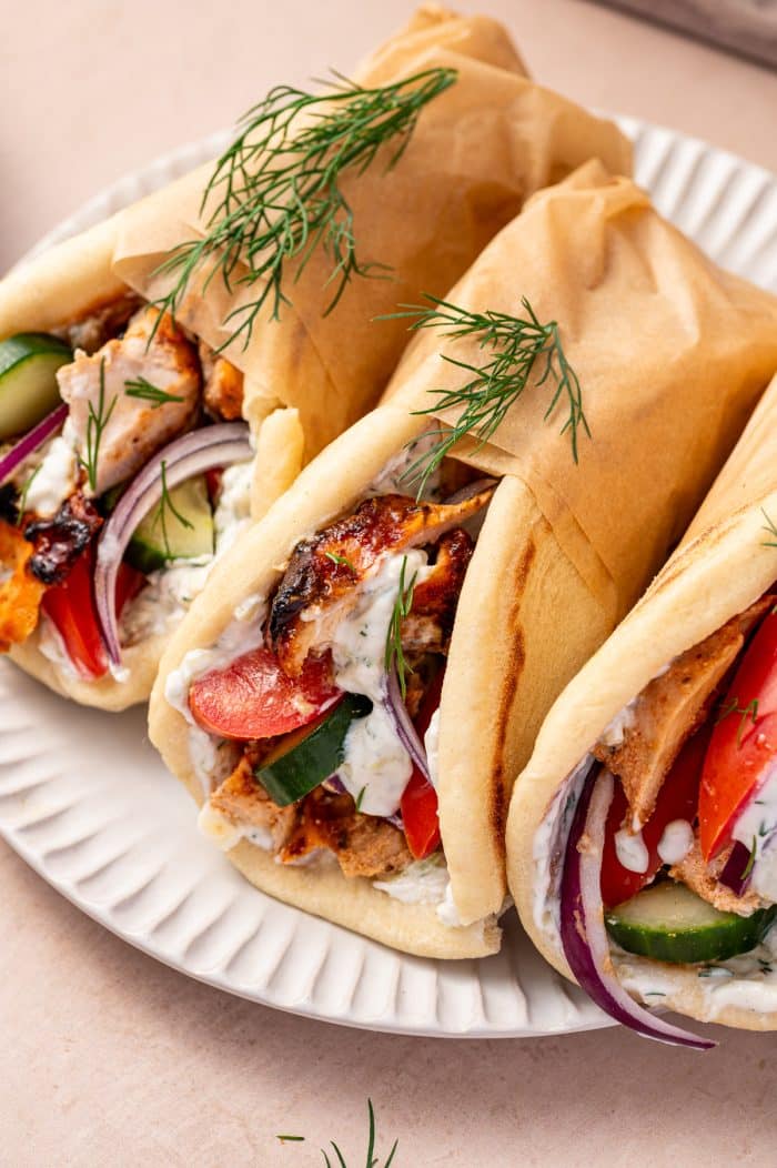 Close-up of three chicken gyro pitas on a white fluted plate, filled with sliced chicken gyro meat, white tzatziki sauce, red tomato slices, cucumber pieces, thin red onion slices, and fresh dill, with the top half of each pita wrapped in brown parchment paper.