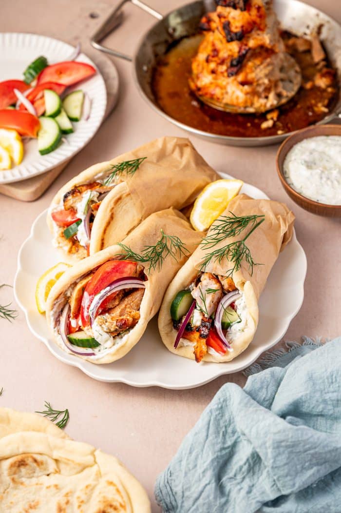 Three homemade chicken gyro wraps on a white scalloped plate, filled with sliced chicken gyro meat, tzatziki, tomatoes, cucumbers, red onion, and fresh dill, with lemon wedges, a bowl of tzatziki, sliced vegetables on a white plate, warm pita bread, a silver pan of roasted chicken gyro meat, and a light blue napkin in the background.