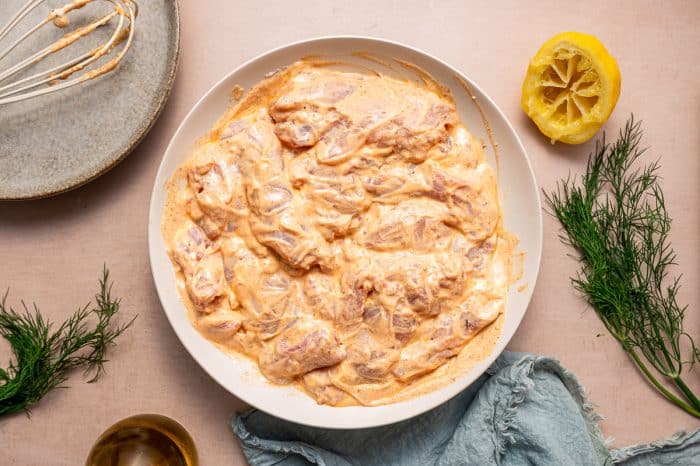 Overhead view of raw chicken thighs coated in creamy orange yogurt marinade in a white shallow bowl, with a whisk on a gray plate, a squeezed lemon half, fresh dill, olive oil, and a blue napkin on a light pink surface.