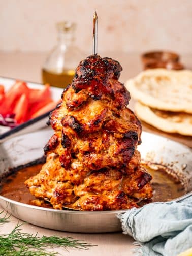 Vertical close-up of roasted chicken gyro meat stacked on a metal spit in a silver pan, with deep golden orange chicken, blackened charred edges, pan juices, warm pita bread, sliced tomatoes, red onion, cucumber, olive oil, fresh dill, and a blue napkin.