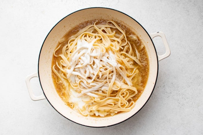 Cooked fettuccini and heavy cream added to a large white skillet filled with a simple sauce of softened aromatics, butter, and pasta water.