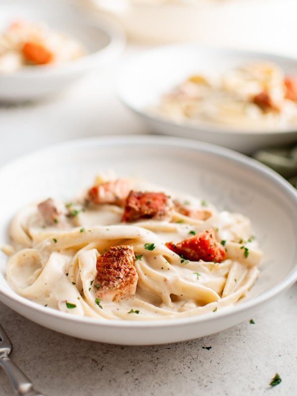 A white bowl filled with fettuccini in cream sauce, topped with crispy Cajun salmon and parsley. Two more bowls appear blurred in the background.