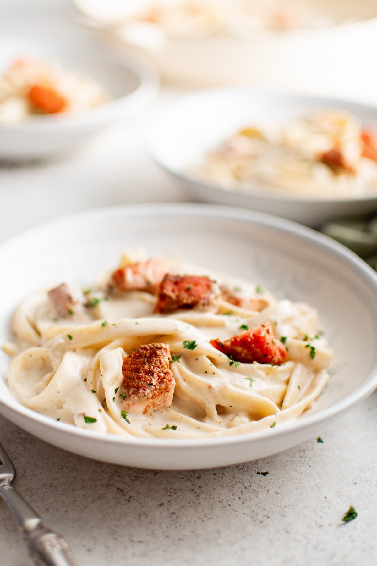 A white bowl filled with fettuccini in cream sauce, topped with crispy Cajun salmon and parsley. Two more bowls appear blurred in the background.