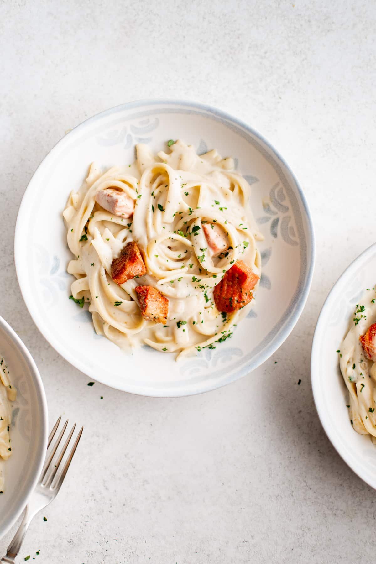 A top-down view of a white bowl filled with creamy fettuccini pasta with salmon pieces.