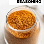 A small glass jar filled with vibrant reddish-orange homemade sazรณn seasoning on a light surface with text overlay "homemade sazon seasoning"