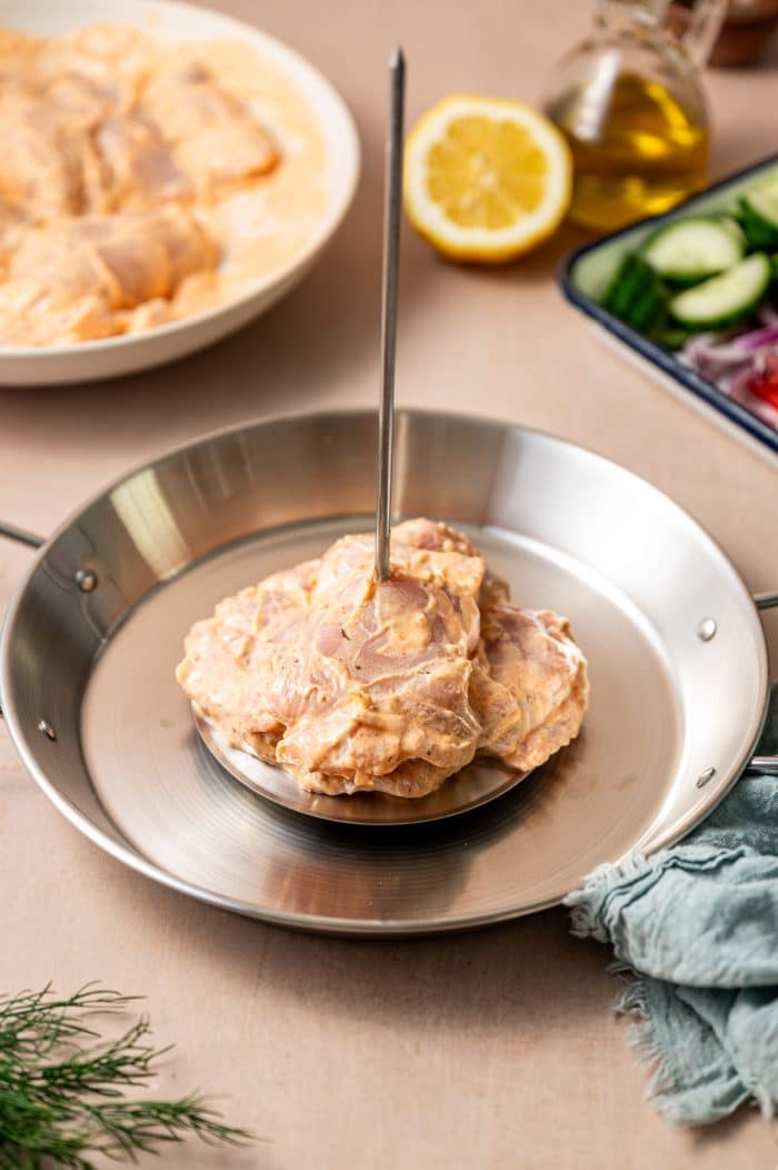 Close-up of marinated chicken thighs stacked on a vertical metal spit in a shiny silver pan, with a bowl of extra marinated chicken, a lemon half, olive oil, sliced cucumbers, red onion, tomatoes, and a blue napkin in the background.