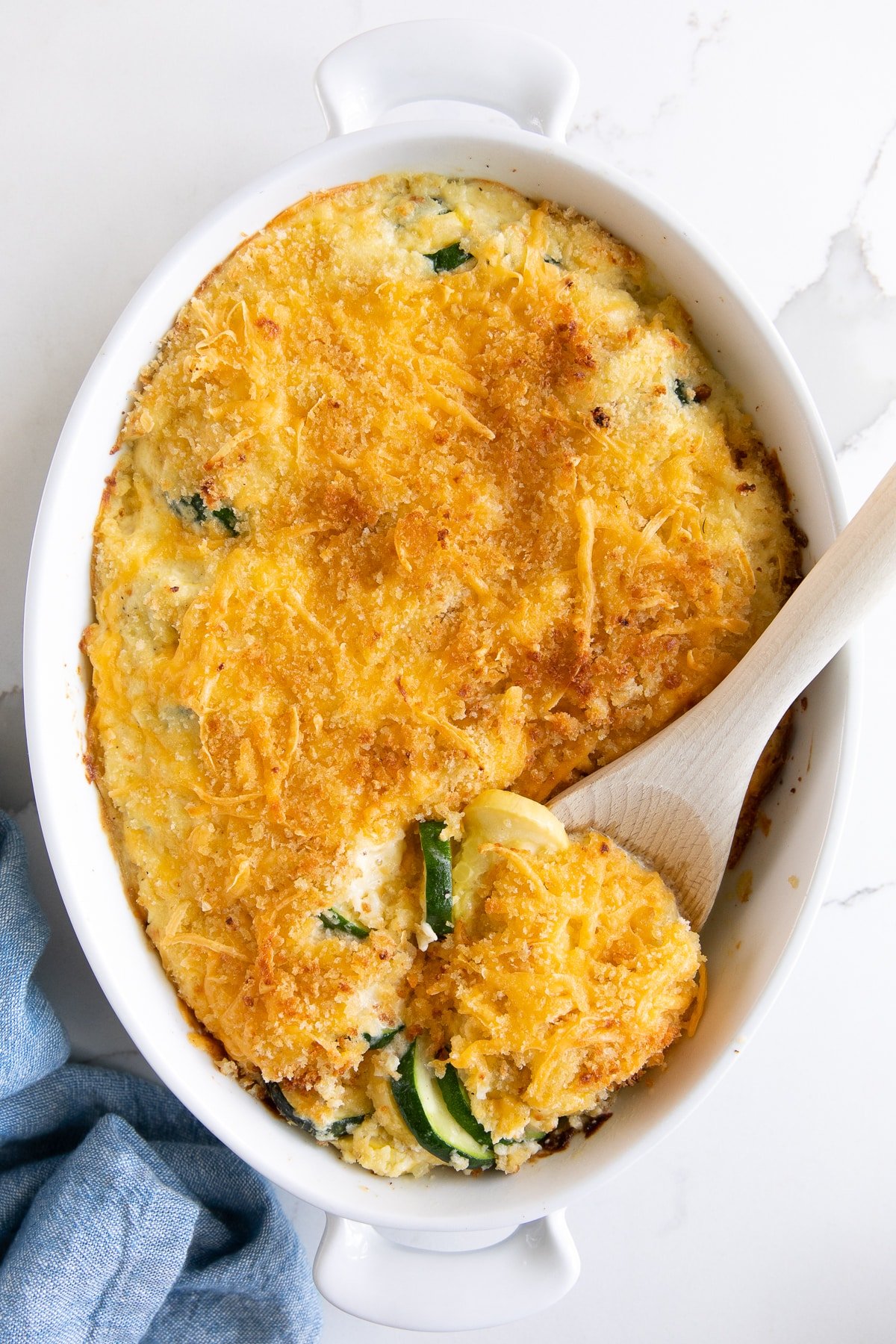 A cheesy, golden-baked zucchini and squash casserole with a crunchy breadcrumb topping, served in a white baking dish with a wooden spoon.