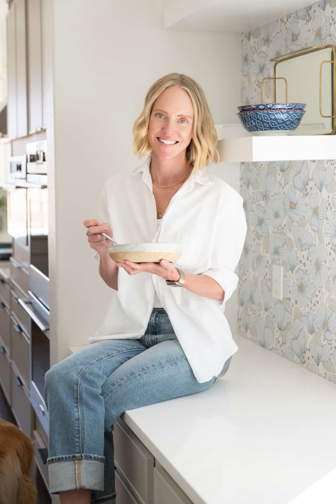 Chef Jessica Randhawa sitting on a kitchen counter holding a bowl of food in her bright home kitchen.