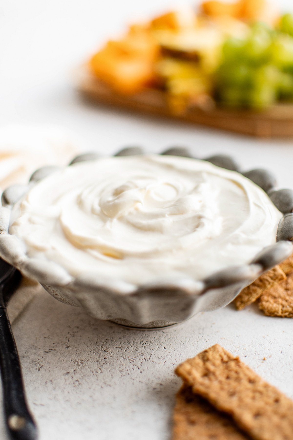 Close-up shot of a decorative bowl filled with cream cheese fruit dip, with graham crackers and a fruit tray in the background.