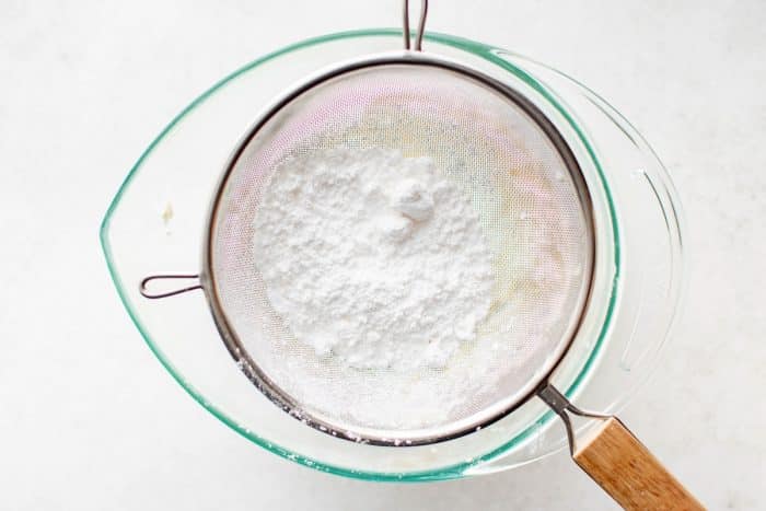 Overhead view of powdered sugar being sifted into a glass mixing bowl with softened cream cheese, ready for blending.