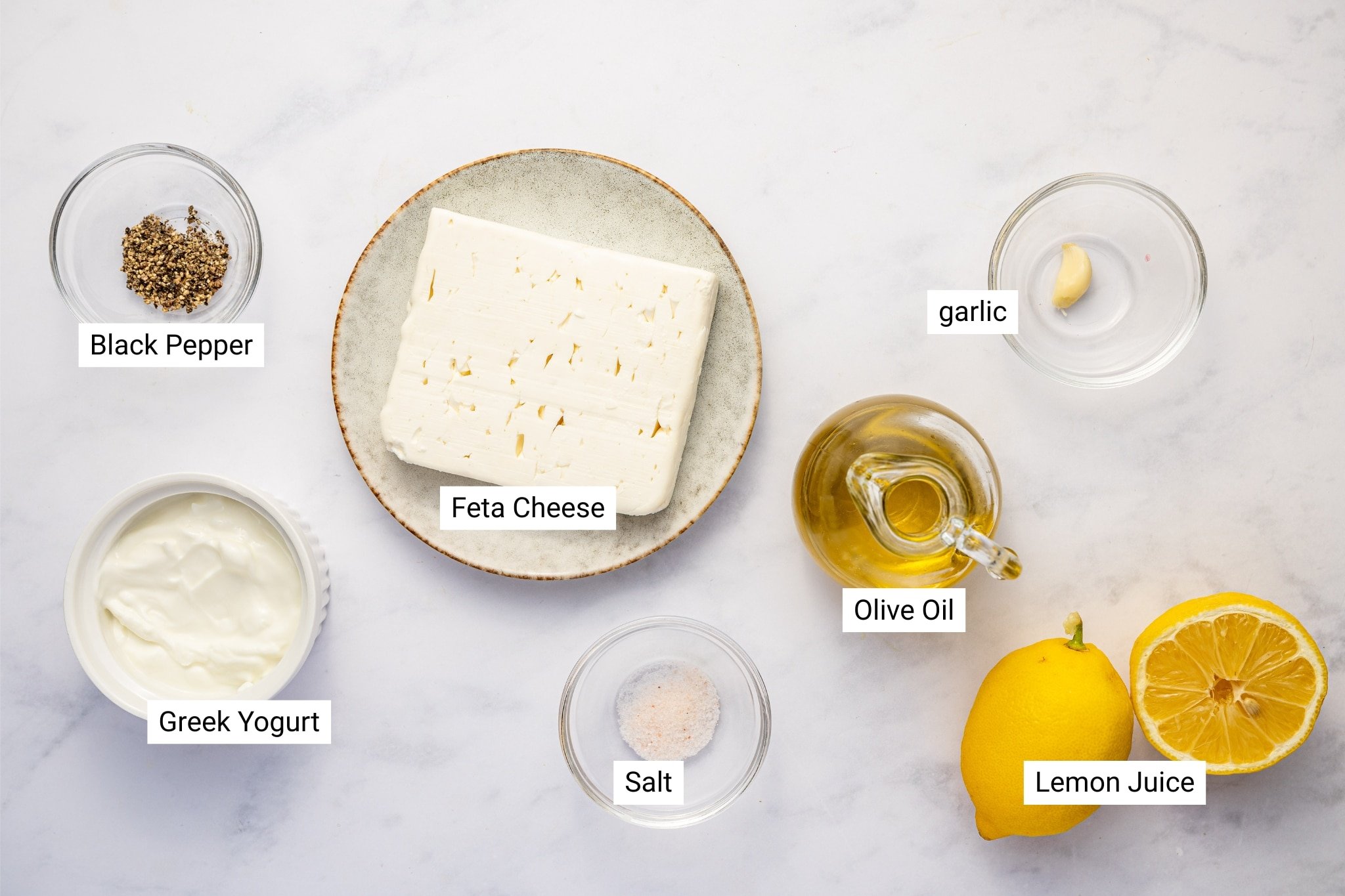 A top-down view of all the ingredients needed to make whipped feta dip. In the center, a block of feta cheese sits on a plate. Surrounding it are small bowls of black pepper, salt, Greek yogurt, olive oil, and a single garlic clove. There is also a halved lemon on the right. The ingredients are clearly labeled and arranged on a white marble countertop.