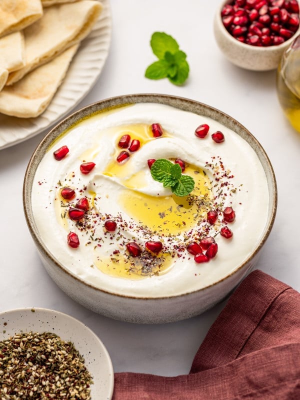 A styled scene of whipped feta dip in a bowl surrounded by pita slices, pomegranate seeds, fresh mint, and za’atar.