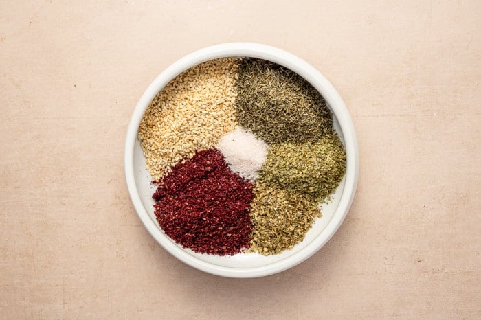 Individual za’atar seasoning ingredients arranged in a white bowl: sesame seeds, thyme, oregano, marjoram, sumac, and salt.