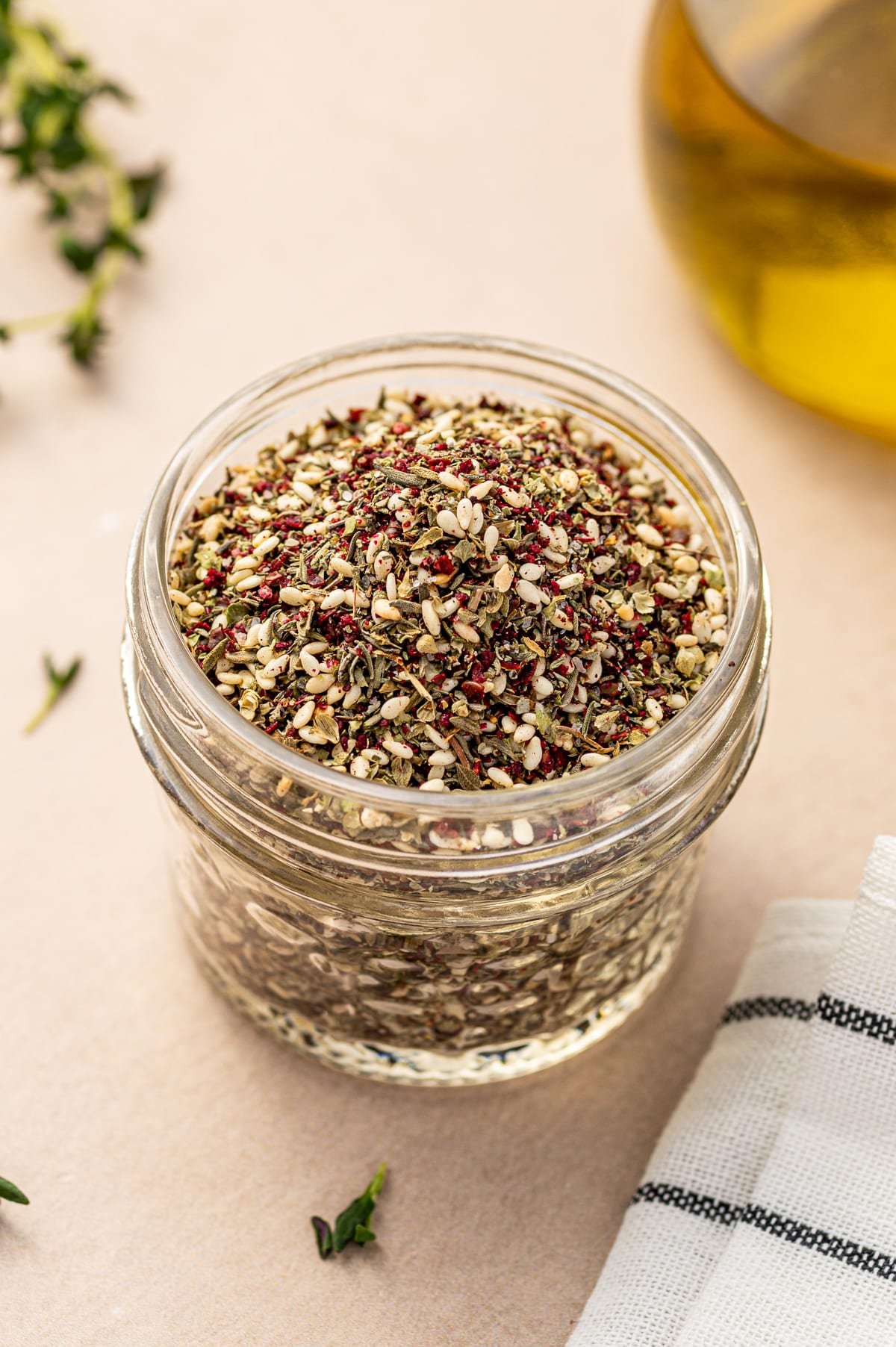 Glass jar filled with za’atar spice blend on a neutral countertop with olive oil in the background.