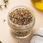 Square image of a a glass jar of za’atar seasoning with herbs and olive oil in the background