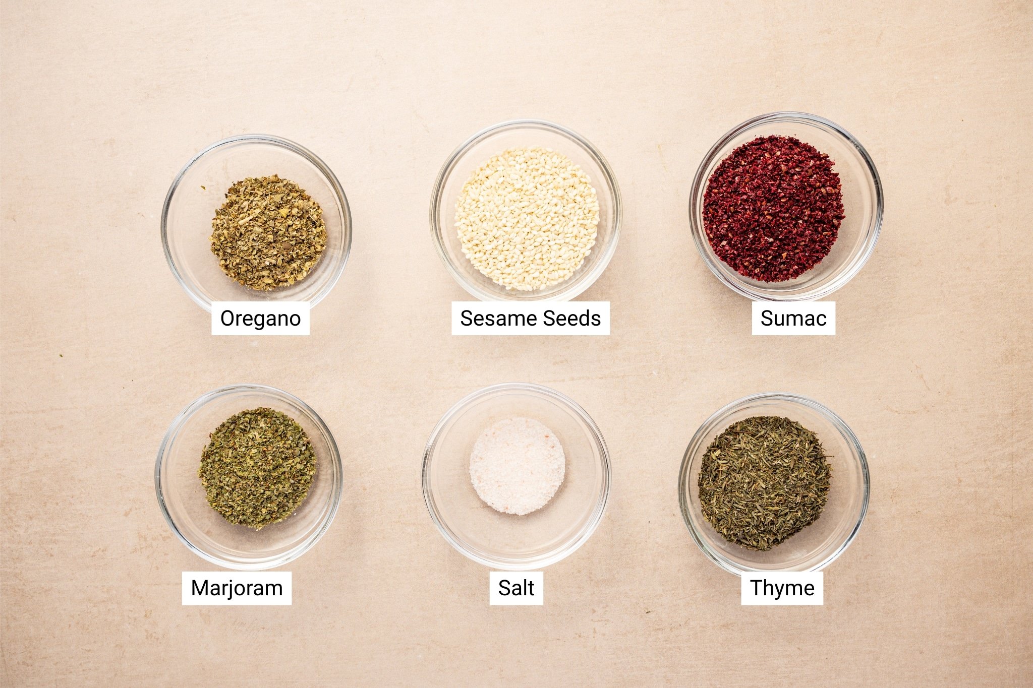 A top-down view of six clear glass bowls, each filled with a different za’atar seasoning ingredient—oregano, sesame seeds, sumac, marjoram, salt, and thyme—arranged in two rows on a light beige background. Each bowl is labeled for clarity.
