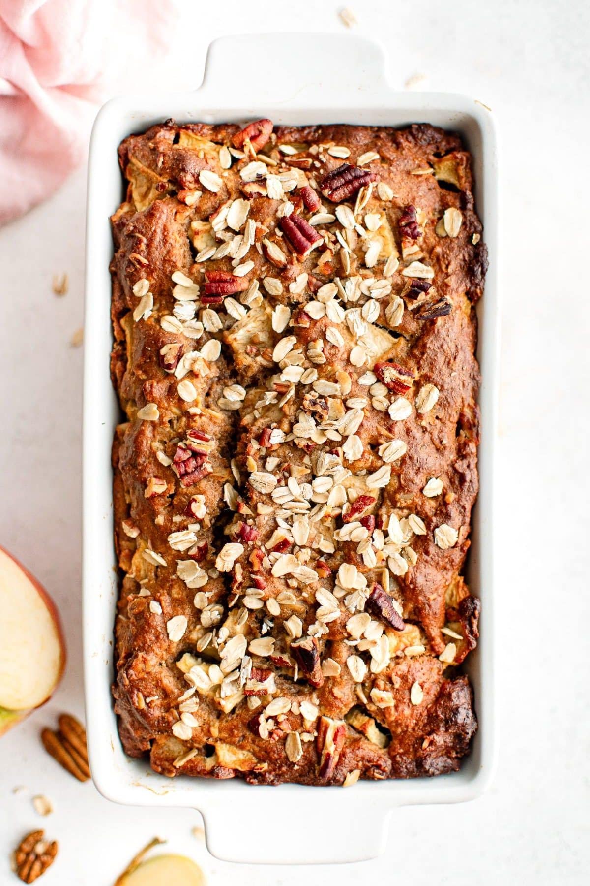 A golden-brown apple cinnamon bread topped with oats and chopped pecans, shown in a white ceramic loaf pan with sliced apples and a pink linen nearby.
